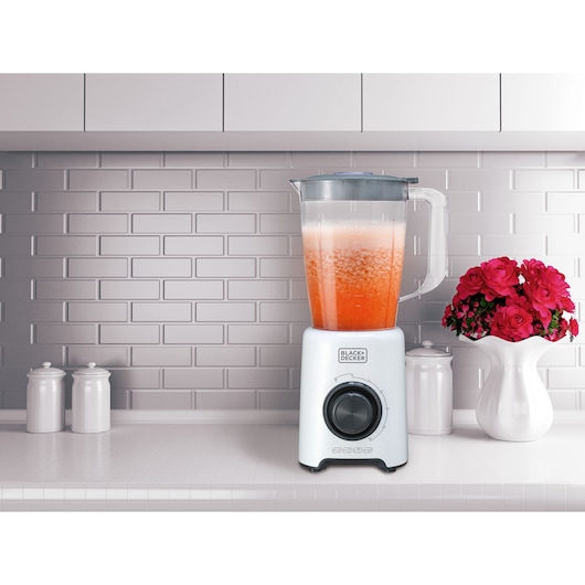 A BLACK+DECKER blender with a transparent pitcher containing a red and orange mixture sits on a kitchen counter. The background features a tiled wall, white cabinetry, two white containers, and a vase with pink flowers.