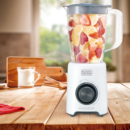 A BLACK+DECKER blender filled with cut fruit sits on a wooden kitchen countertop. Next to the blender is a glass cup of milk and a wooden cutting board. The background is softly blurred, suggesting a relaxed kitchen atmosphere.
