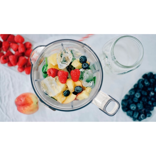 Top view of a blender jug containing ice, pineapple pieces, spinach, blueberries, and raspberries, with ingredients like blueberries, raspberries, and an apple arranged around it on a white surface.
