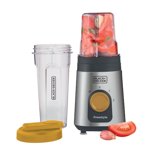 A BLACK+DECKER personal blender with a metallic base, yellow control dial, a blending cup filled with chopped tomatoes, a second empty blending cup, a yellow lid, and pieces of tomato and garlic beside the blender.