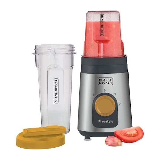 A BLACK+DECKER blender with a gray and black base, a yellow dial, and labeled 'Freestyle'. One blending cup is filled with a red smoothie and attached to the base. An empty cup and a yellow lid are placed next to the blender. There is a piece of tomato and a garlic clove in front of the blender.