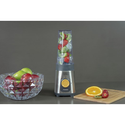 A BLACK+DECKER blender filled with strawberries and kiwi sits on a counter. To the left, there is a glass bowl filled with green and red apples. To the right, a wooden cutting board holds half an orange, a sliced kiwi, and a strawberry. The background is plain gray.