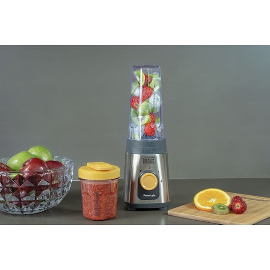 A BLACK+DECKER blender with fruits inside, next to a jar containing blended fruit, a bowl of apples, and a wooden board with sliced orange, kiwi, and strawberry on a gray surface.
