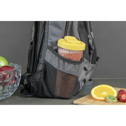 A portable blender bottle with a yellow lid is placed in the mesh side pocket of a grey backpack. Fresh fruits including an orange slice, kiwi, and strawberries are on a wooden board, and apples are in a glass bowl on a table nearby.