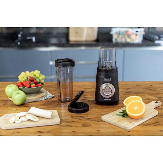 A BLACK+DECKER blender with two portable cups and a lid is displayed on a wooden kitchen counter. Surrounding the blender are plates and boards with fresh fruits including apples, grapes, strawberries, a banana, and sliced oranges.