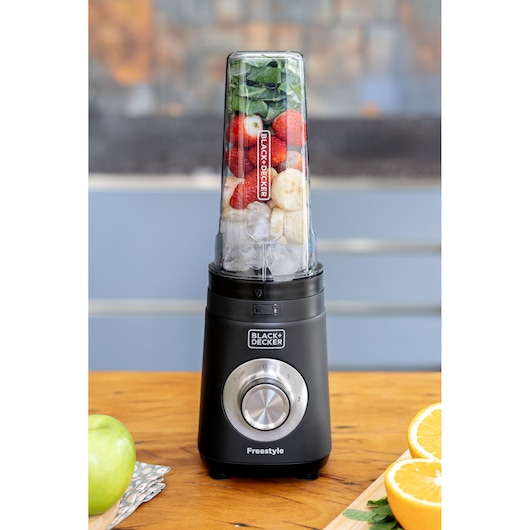A BLACK+DECKER Freestyle blender with a transparent container filled with spinach, strawberries, banana slices, and ice, placed on a wooden surface. Sliced oranges and a green apple are visible nearby.