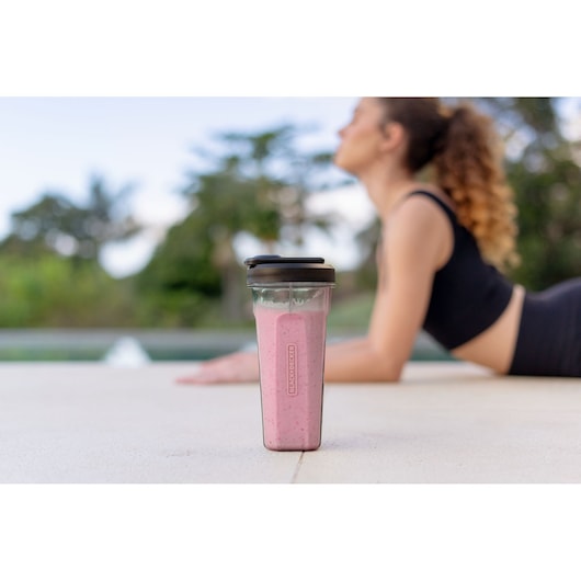 A BLACK+DECKER blender bottle filled with a pink smoothie is placed on the ground outdoors, with a person in athletic clothing blurred in the background near a pool.