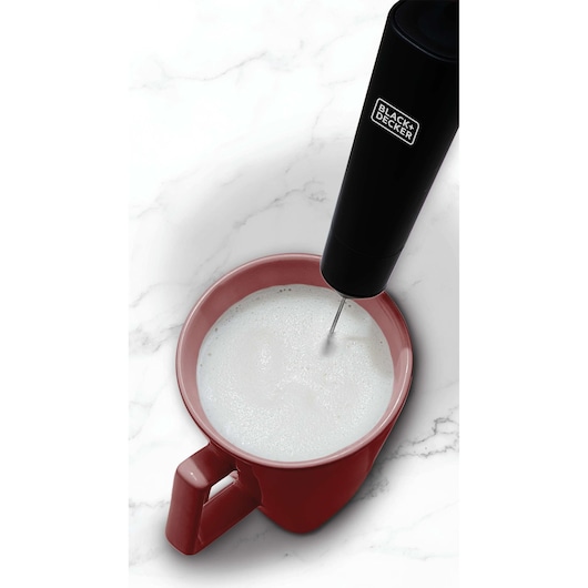 A BLACK+DECKER handheld milk frother being used to froth milk in a red ceramic mug on a white marble surface.