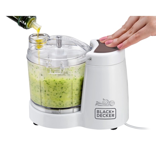 BLACKDECKER MP120 food processor with clear bowl, green mixture inside, oil being poured, hand pressing lid, front angle.