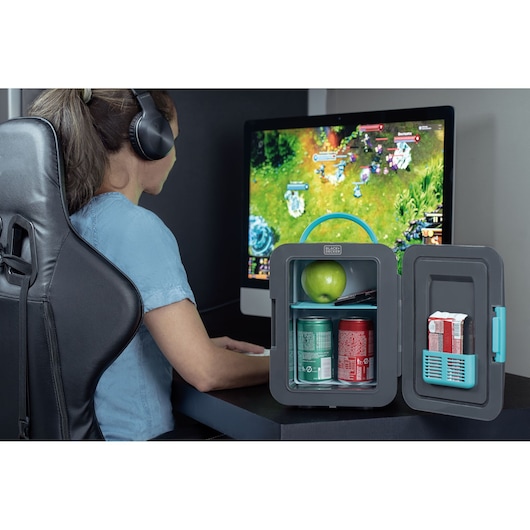 BLACKDECKER MR60 compact mini fridge on desk open with drinks, apple, and snacks, next to person gaming on computer.