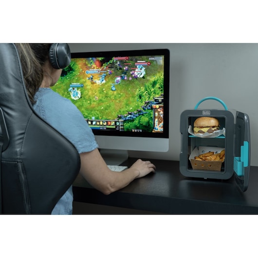 Photo of BLACKDECKER MR60 mini fridge on desk with cheeseburger and fries inside, next to person gaming at computer.