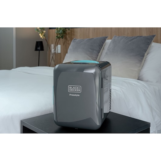 A BLACK+DECKER Freestyle compact portable appliance is placed on a black table next to a neatly made bed with white sheets and grey pillows in a modern bedroom.