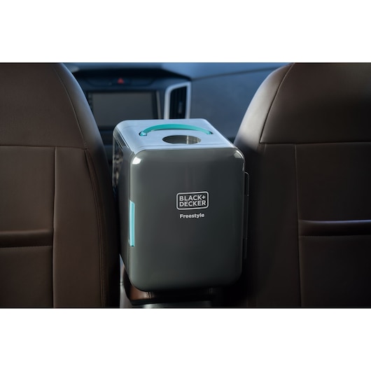 A BLACK+DECKER Freestyle portable refrigerator is shown placed between the front seats of a car. The compact appliance has a dark grey color with a turquoise accent and is designed for travel or on-the-go cooling.