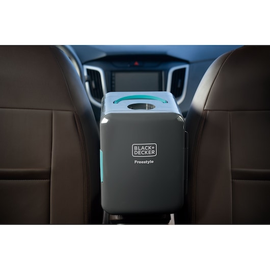 A BLACK+DECKER Freestyle portable cooler is shown placed between two front seats inside a car. The unit is grey with turquoise accents and has a circular opening on the top.