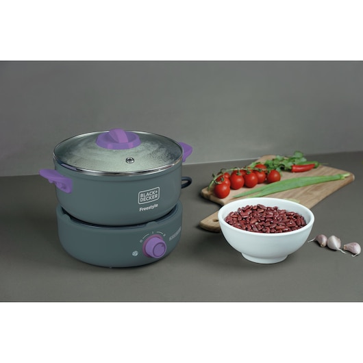 A BLACK+DECKER Freestyle electric cooker with purple handles and a glass lid is placed on a kitchen counter. Next to it is a white bowl filled with red beans, three cloves of garlic, and a wooden cutting board with cherry tomatoes, a red chili, green onion, and herbs.