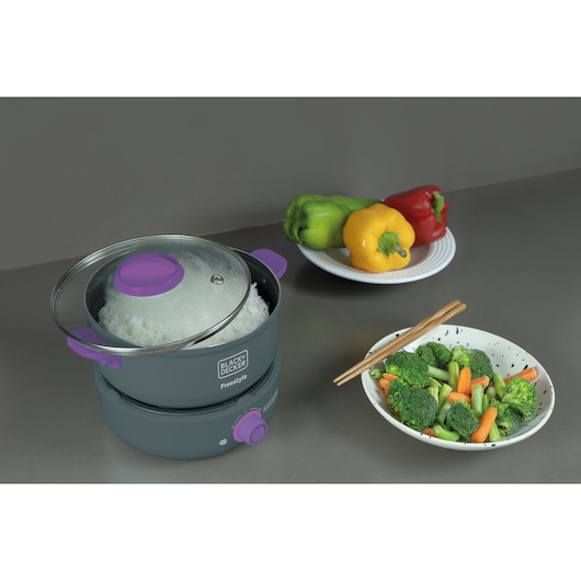 A BLACK+DECKER Freestyle electric rice cooker with purple accents on a grey countertop. The cooker contains cooked rice and is placed next to a plate of bell peppers and a bowl of steamed vegetables with chopsticks.