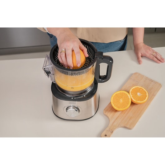 A person uses a BLACK+DECKER electric juicer to squeeze an orange, with fresh orange halves placed on a wooden cutting board next to the juicer.