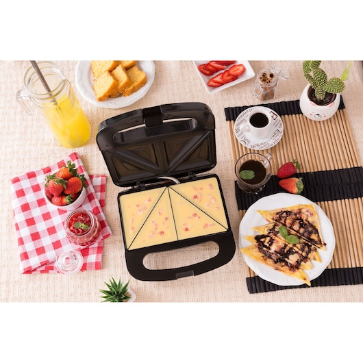 A BLACK+DECKER sandwich maker model SM800 is open on a table, filled with a baking mixture. Surrounding it are plates of strawberries, cake, sliced strawberries, a glass jar of juice, coffee cups, a plate of finished triangle sandwiches with chocolate drizzle and mint leaves, and assorted breakfast items.