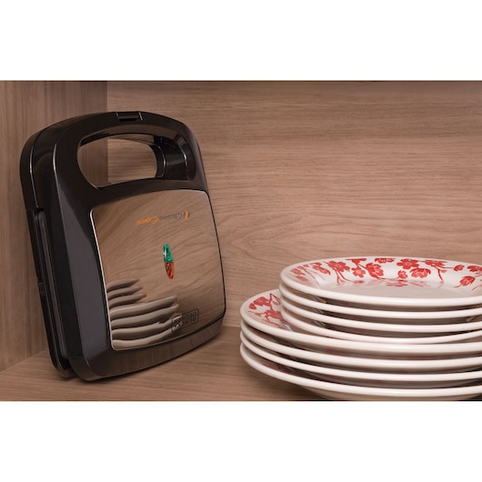 A BLACKDECKER sandwich maker model SM800 with a metallic finish is placed upright on a wooden shelf next to a stack of white plates with red floral patterns.