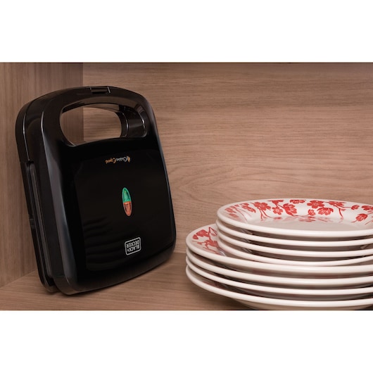 A BLACK+DECKER sandwich maker placed vertically in a wooden kitchen cabinet next to a stack of white plates with red floral designs.