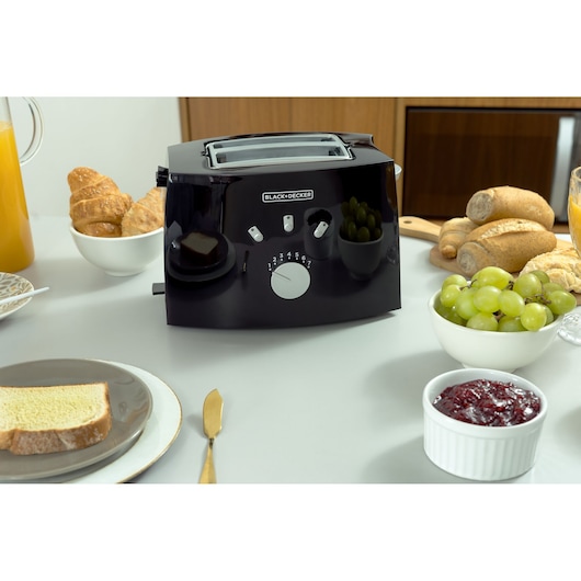 BLACK+DECKER TO810 black toaster on kitchen table, surrounded by bread, grapes, orange juice, pastries, and jam.