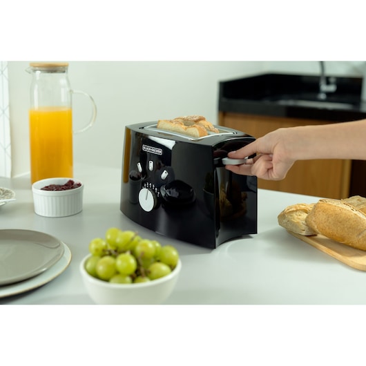 BLACK+DECKER TO810 black toaster on kitchen counter with bread inside, hand pressing down lever, fruit and juice nearby.