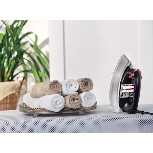A BLACK+DECKER electric iron standing upright on a patterned ironing board next to a decorative tray holding rolled towels in beige and white colors. There is a green plant in the background.