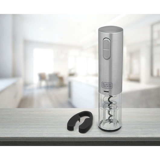 A Black+Decker electric wine opener with a stainless steel finish, shown on a countertop next to a black foil cutter. The background features a modern, blurred kitchen scene.