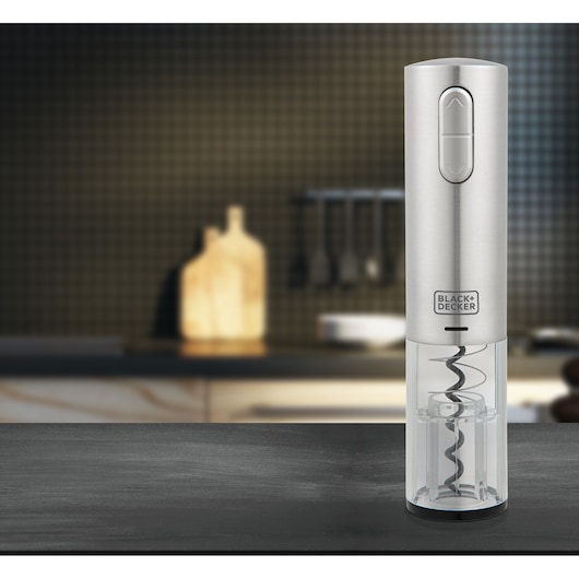 A BLACK+DECKER electric wine bottle opener with a stainless steel finish is shown standing upright on a kitchen counter. The opener features a transparent lower section displaying the corkscrew mechanism. The background is a modern kitchen with blurred utensils and a cutting board.
