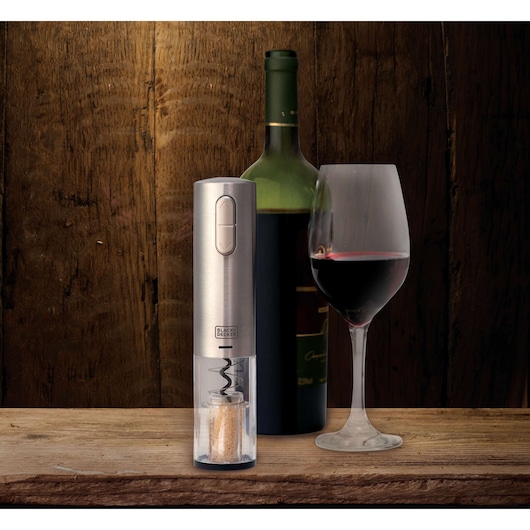 A BLACK+DECKER electric wine opener is displayed on a wooden surface next to a bottle of wine and a glass filled with red wine. The background is made of wood.