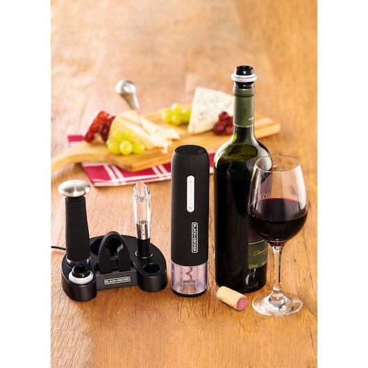 A BLACK+DECKER wine accessory set displayed on a wooden table, including an electric wine opener, aerator, vacuum pump, foil cutter, and charging base. There is a bottle of red wine, a glass of wine, a cork, and a cheese platter with grapes in the background.