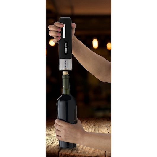 A person uses a BLACK+DECKER branded electric wine opener to open a bottle of wine, holding the opener vertically on the cork and securing the bottle with the other hand. The scene is set on a wooden table with blurred warm lights in the background.